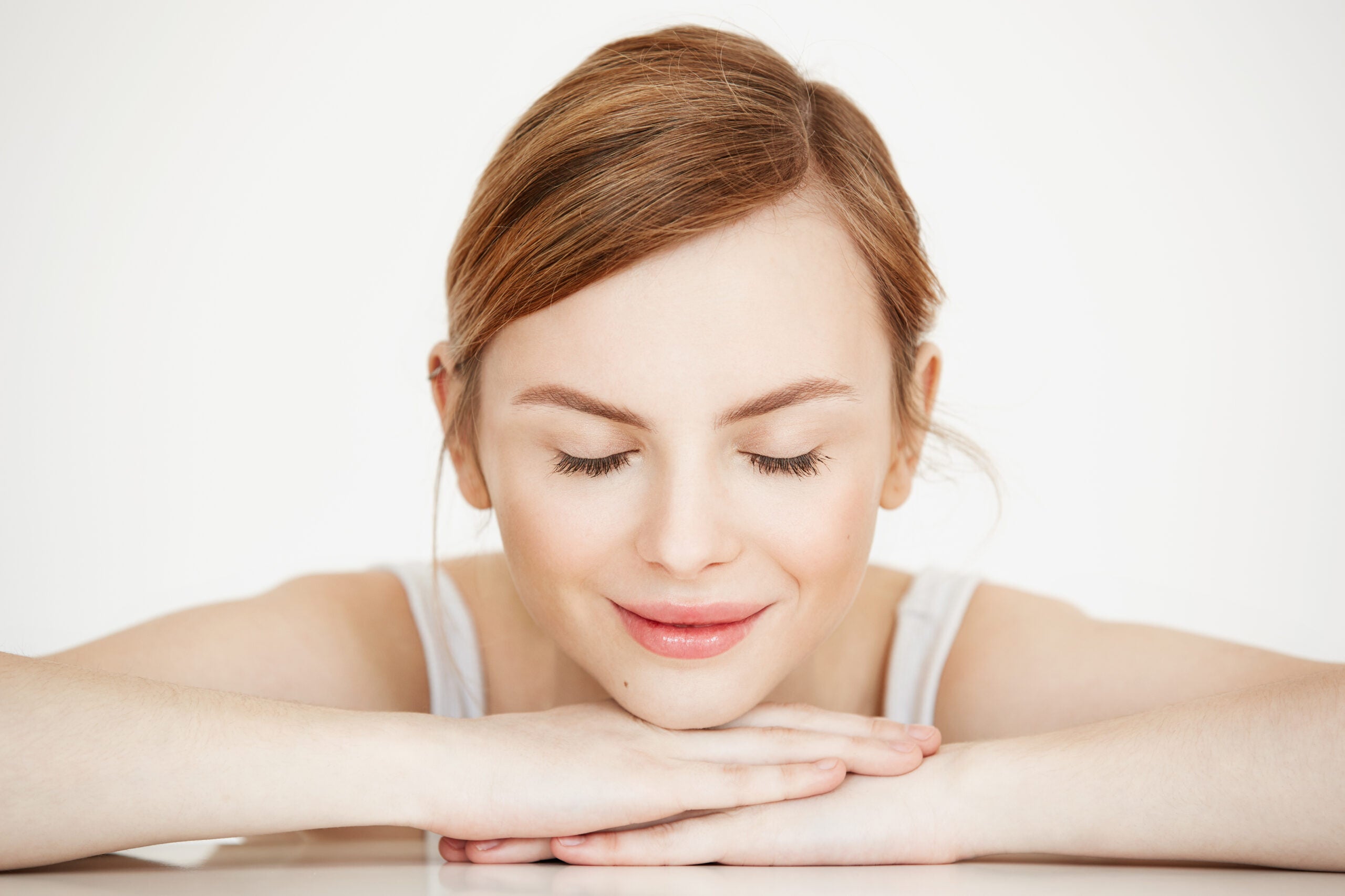 Young beautiful girl with perfect clean skin smiling with closed eyes sitting at table over white background. Beauty spa and cosmetology.